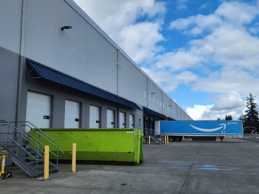 Large lime green roll-off dumpster rental parked at an Auburn, WA warehouse loading dock next to an Amazon Prime semi-truck.